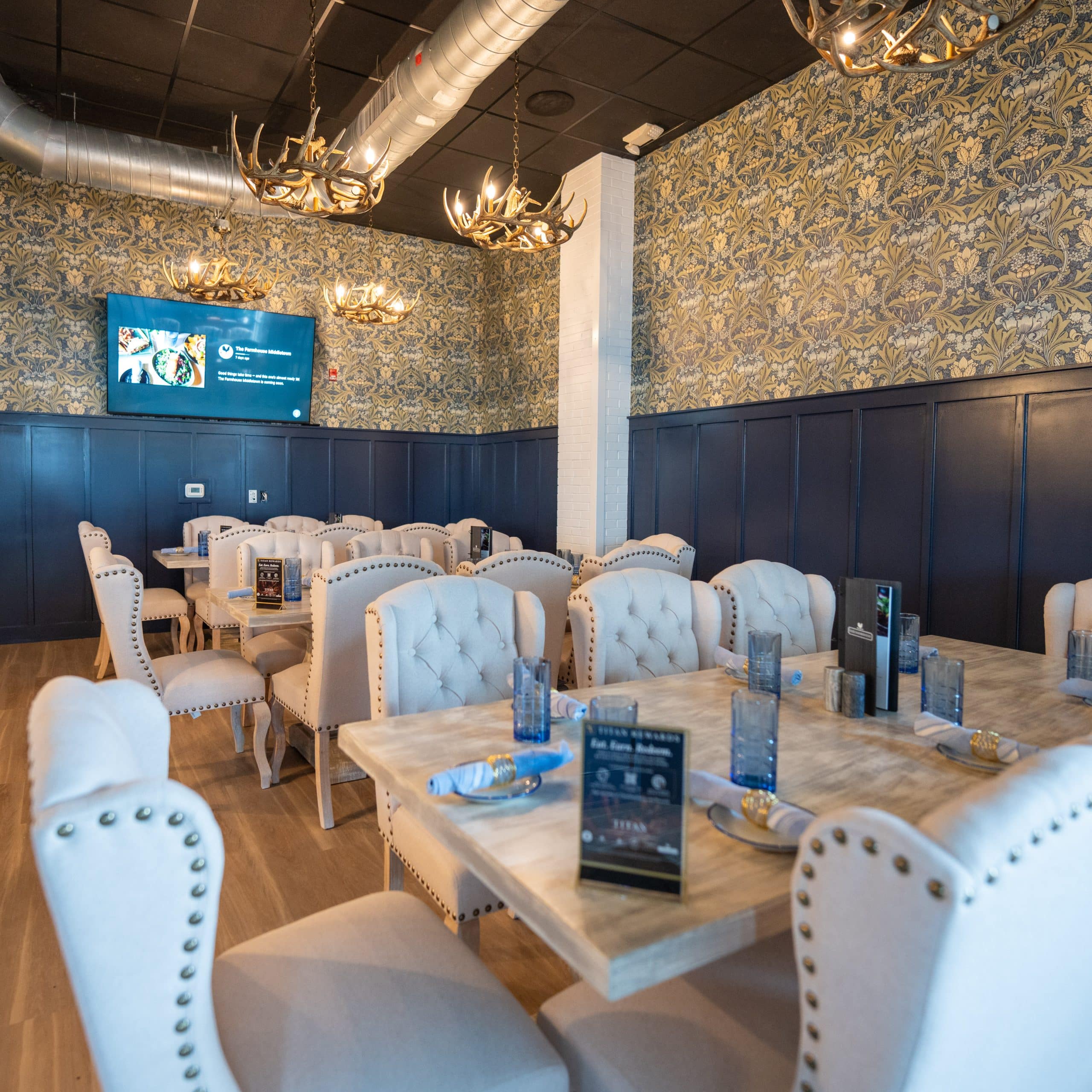 Dining room in a restaurant with tufted white chairs, blue wainscoting, and antler chandeliers against patterned wallpaper.